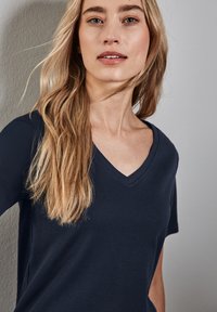 Námořnická modrá v-neck tričko z měkké látky, s krátkými rukávy a uvolněným střihem. Jemná textura zvyšuje vizuální zajímavost.