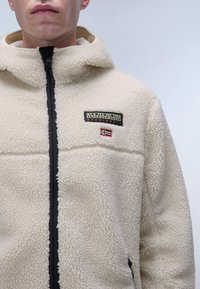 Beige fleece jacket with a front zipper, textured fabric, black accents, and embroidered logo. Features a hood and side pocket.