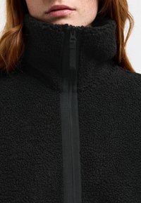 Svart fleecejacka med hög krage, som har full dragkedja och texturerad yta. Designen prioriterar värme och komfort.