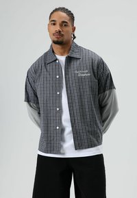 Kortemouw overhemd met ruitpatroon in grijs en zwart met witte knopen, met de tekst "Can't crush greatness" geborduurd op de borst.