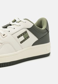 Zapatilla blanca y verde oliva con parte superior de cuero, cuello acolchado, suela de goma y un parche rectangular con el texto "TOMMY JEANS".