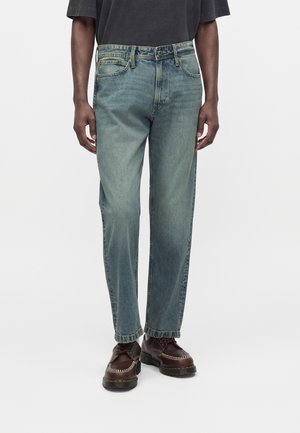 Personne portant un jean bleu délavé et des chaussures en cuir marron foncé, debout devant un fond clair uni.