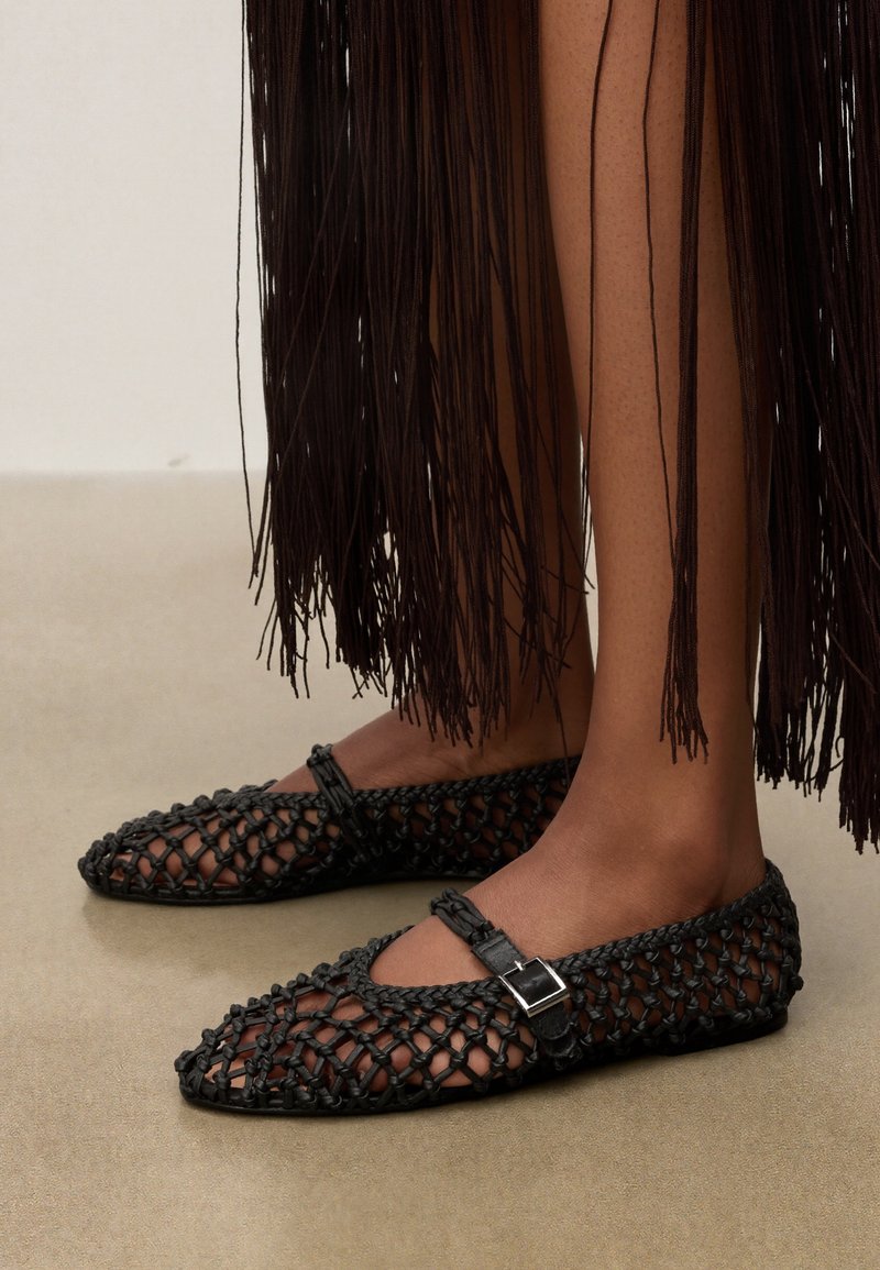 Ballerines Mary Jane noires tissées avec des brides à boucle portées avec une longue jupe marron foncé à franges, montrant des jambes et des pieds nus sur un sol neutre.