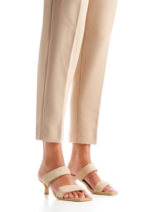Beige suede heeled sandals with two wide straps over the foot and a slender heel. Paired with cream-colored tapered pants.