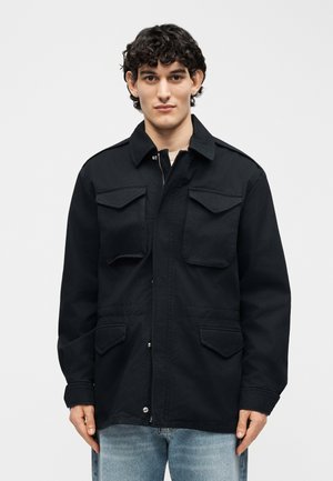 Mladý muž s kučeravými tmavými vlasmi v čiernej bunde so štyrmi vreckami s chlopňami a svetlomodrými džínsami, stojaci pred jednoduchým pozadím.