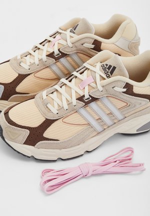 Beżowo-brązowe sneakersy Adidas z kremowymi sznurowadłami, trzema przezroczystymi bocznymi paskami oraz dodatkowymi różowymi sznurowadłami obok nich.