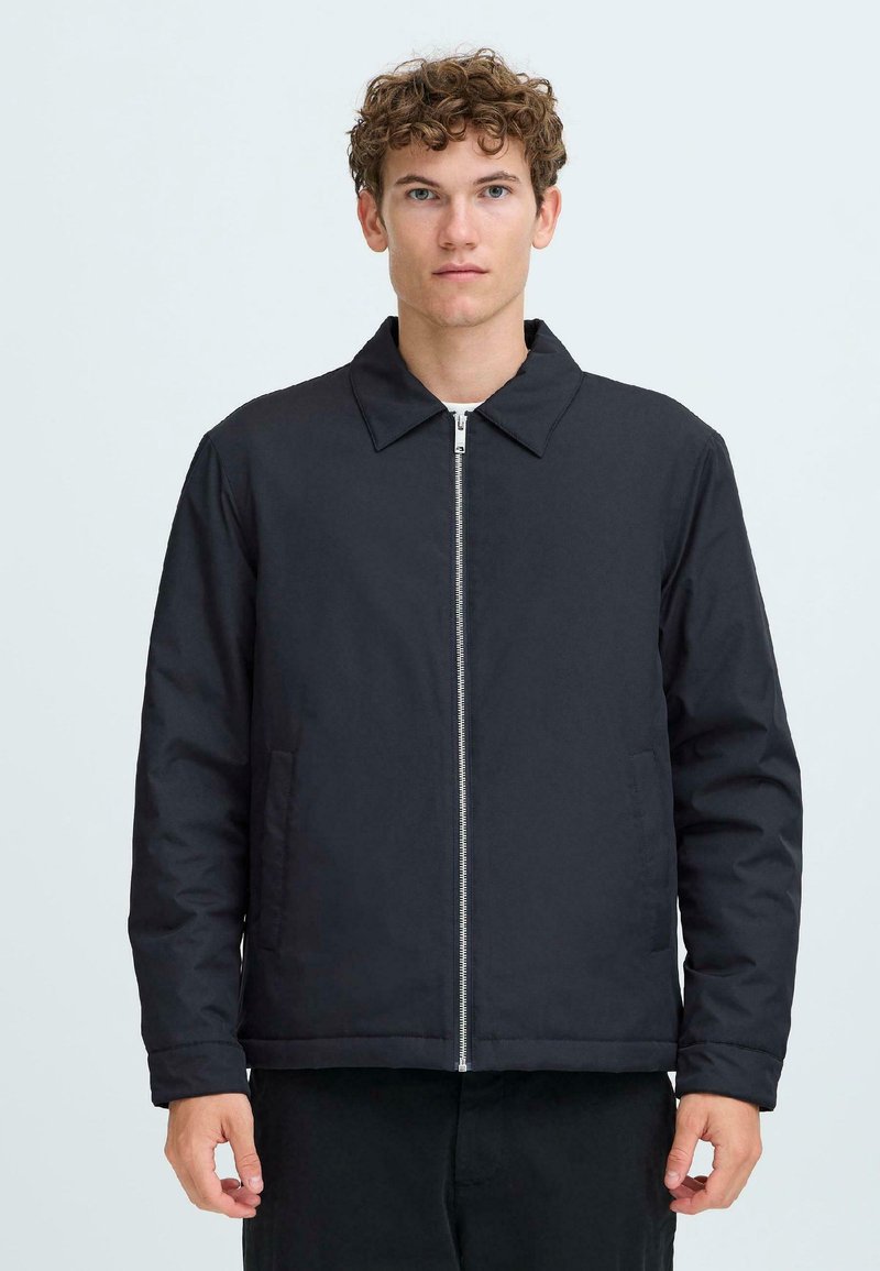 Young man with curly hair wearing a black zip-up jacket with collar, standing against a plain light background.