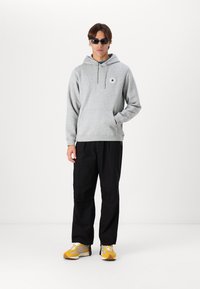 Grå hettegenser med frontlomme, blå detalj og logo, kombinert med svarte bukser og gule sneakers med grå detaljer.