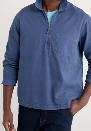 Mężczyzna ubrany w niebieską bluzę na suwak do połowy piersi z kieszenią na piersi oraz jasnoniebieską koszulę pod spodem, z jedną ręką w kieszeni ciemnoniebieskich dżinsów.