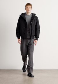 Veste noire à capuche avec des fermetures à pression, chemise à manches longues grise, pantalon rayé et chaussures noires à enfiler. Design minimal et texture douce.