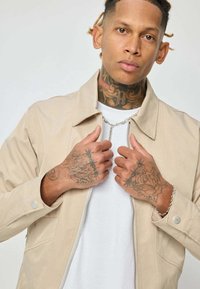 Un homme avec des tatouages au cou et sur les mains porte une veste beige par-dessus une chemise blanche, tenant le col de la veste avec les deux mains.