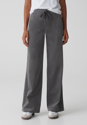 Personne portant un pantalon large en velours côtelé gris avec taille à cordon et des baskets blanches, debout sur un fond uni.