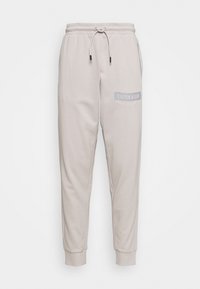 Light beige cotton joggers with a drawstring waist, tapered legs, and a soft texture. Features a gray "CALVIN KLEIN" logo on the left side.