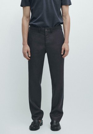 Persona che indossa pantaloni neri eleganti, mocassini neri e una maglietta grigio scuro, in piedi davanti a uno sfondo chiaro semplice.