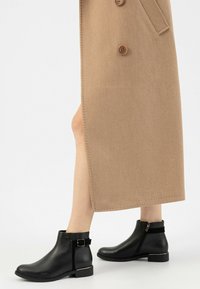 Stivaletti neri in pelle con zip laterale, dettaglio con fibbia e tacco basso a blocco, abbinati a un cappotto di lana beige. Texture liscia.