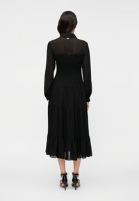 Čierne šaty s priliehavým žabičkovaným živôtikom, dlhými rukávmi a vrstvenou, rozšírenou sukňou s volánovými akcentmi. Priehľadný zadný detail s golierom.