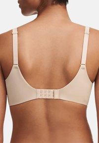 Beżowy, bezszwowy biustonosz z regulowanymi ramiączkami i zapięciem na haftkę z tyłu. Gładka tekstura, minimalistyczny design i zaokrąglony kształt.