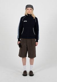 Une femme se tient face à nous, portant un pull zippé bleu marine avec des rayures bleu clair, un short marron au genou, un béret gris, des chaussettes blanches et des chaussures foncées.