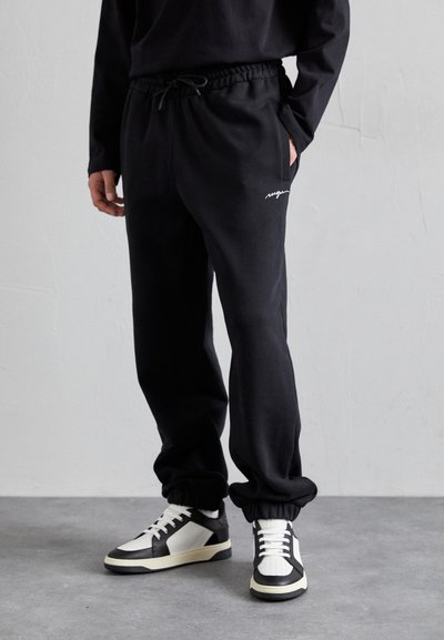 Pantalons de jogging noirs avec une taille élastique, des poches latérales, des chevilles resserrées et un discret logo blanc, accompagnés de baskets noires et blanches.