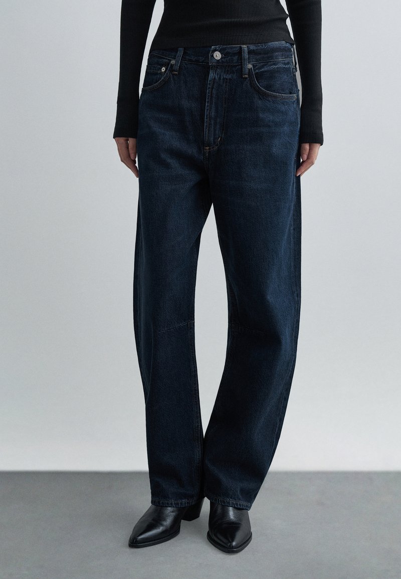 Sötétkék, széles szárú farmernadrág farmer anyagból, gombos sliccel, elülső zsebekkel és enyhe fényes felülettel. Fekete hegyes sarkú cipővel párosítva.