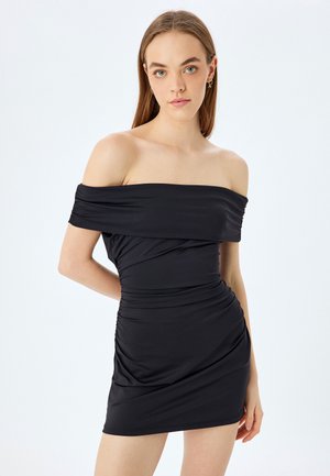 Sort off-shoulder kjole lavet af strækbart stof. Den har indsamlet tekstur og rynkede detaljer langs kroppen og slutter lige over knæet.