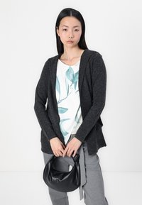 Charcoal gray cardigan with an open front, worn over a white top featuring a teal leaf print. Model holds a black hobo bag, gray pants visible.