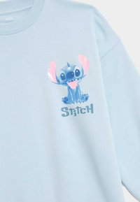 Felpa azzurra chiara con stampa grafica di un personaggio dei cartoni animati con grandi orecchie, accompagnata dal testo "Stitch" in lettere grasse.
