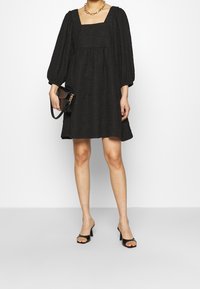 Svart texturerad klänning med fyrkantig halsringning och puffärmar. Bärs med en svart handväska och öppna klackar. Enkel och avslappnad design.