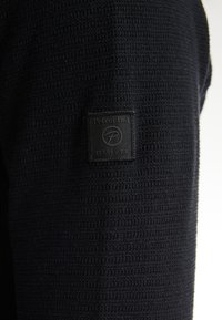Black knit shirt featuring a textured pattern and a woven label with "Petrol Ind. EST. 1989 U.S.A." stitched on the left sleeve.
