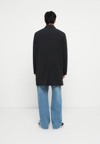 Μαύρο μακρύ σακάκι με γιακά, με ευθεία γραμμή και πλευρικές σχισμές. Συνδυασμένο με ανοιχτό μπλε παντελόνι με φαρδιά πόδια και μαύρα παπούτσια.