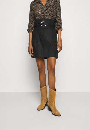 Jupe en denim noir avec une large ceinture ornée d'une boucle en argent circulaire, associée à des bottes en daim beige à hauteur de genou et un haut à motifs.