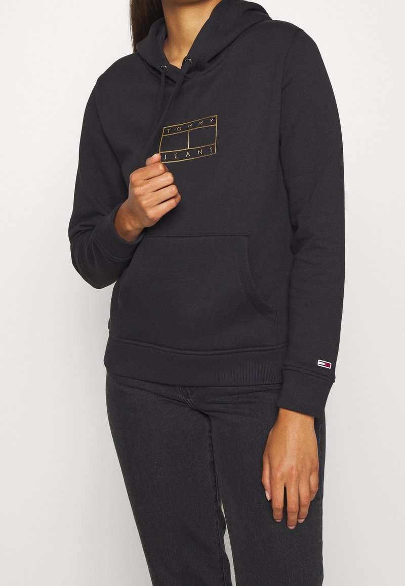 Mujer que lleva una sudadera negra con el logo dorado de "Tommy Jeans" en el pecho y una pequeña bandera de Tommy Hilfiger en el puño izquierdo de la manga.