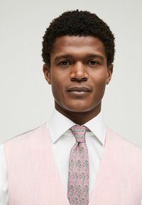 Uomo con capelli corti e ricci indossa una camicia bianca, una cravatta a fantasia rosa e un gilet rosa chiaro su uno sfondo semplice e chiaro.