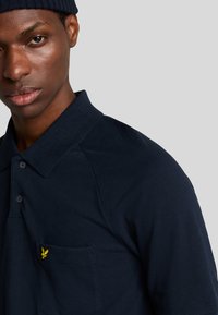 Marine polo-shirt i struktureret stof, med klassisk krave og en enkelt brystlomme med et gult emblem. To knapper ved halsen.