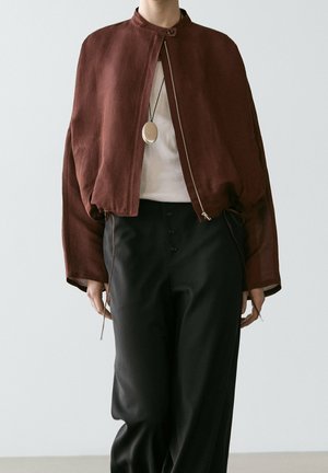 Personne portant une veste courte marron zippée, un haut blanc, un pantalon noir taille haute et un collier avec pendentif rond sur un fond uni.