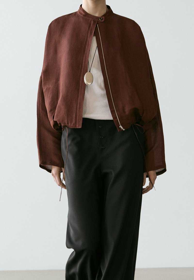 Personne portant une veste courte marron zippée, un haut blanc, un pantalon noir taille haute et un collier avec pendentif rond sur un fond uni.