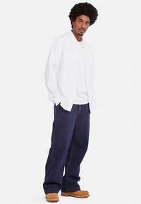 Bijela košulja na kopčanje preko bijele majice, u kombinaciji s tamnoplavim širokim hlačama i smeđim čizmama. Tekstura je glatka; hlače imaju vidljive šavove.