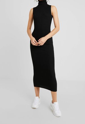 Vrouw draagt een mouwloze zwarte geribde midi coltrui jurk gecombineerd met witte chunky sneakers, staand met de handen gevouwen voor zich.