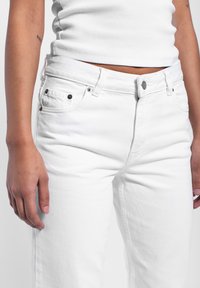 Jeans de mezclilla blanca con un diseño de corte recto, que presentan detalles de herrajes metálicos y bolsillos frontales, combinados con una camiseta sin mangas de canalé blanca.