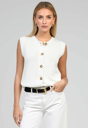 Femme portant un gilet sans manches en tricot blanc à boutons, avec de grands boutons marron, un pantalon blanc, une ceinture noire à boucle dorée et des boucles d'oreilles créoles dorées.