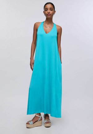 Kobieta w turkusowej sukience maxi bez rękawów i jasnoniebieskich sandałach na koturnie, stojąca na jednolitym jasnym tle.