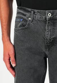 Jeans de mezclilla grises con un ajuste estructurado, bolsillo frontal con costuras azules y herrajes metálicos en la cinturilla. La textura parece suave.