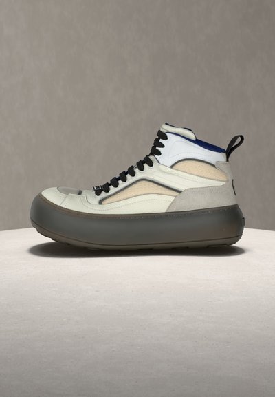 Vysoká teniska s bielymi a béžovými panelmi, čiernymi šnúrkami, modrým vnútrom a hrubou tmavosivou podrážkou na svetlom povrchu proti neutrálnemu pozadiu.