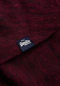 Gros plan sur un tissu tricoté marron avec une petite étiquette bleu marine portant l'inscription "Real SuperDry" et des caractères japonais blancs.