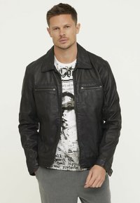 Veste en cuir noir avec un col classique, dotée de poches zippées et d'une texture lisse, associée à un t-shirt graphique blanc en dessous.