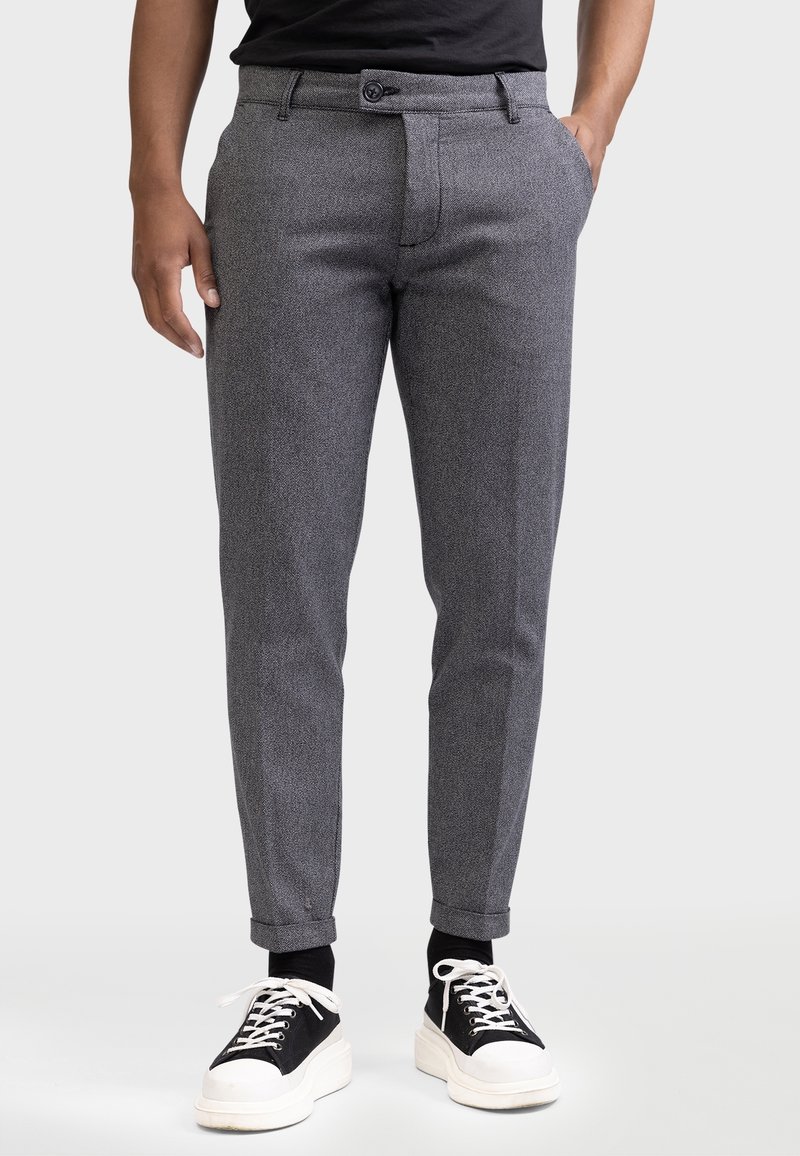 Homme portant un pantalon gris texturé ajusté avec revers, des chaussettes noires, des baskets à lacets noires et blanches, et une chemise noire, debout les mains dans les poches.