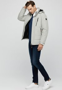 Chaqueta acolchada gris claro con capucha, que presenta bolsillos con cremallera, un forro negro y conjunto a juego con top azul oscuro y vaqueros azules, combinados con zapatillas blancas.