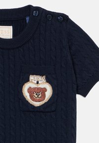 Κοντομάνικο μπλε ναυτικό πλεκτό τοπ με στρογγυλή λαιμόκοψη. Διαθέτει υφή με μοτίβο καλωδίου και κεντημένο πατς με θέμα αρκούδας στο στήθος.