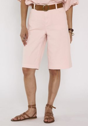 Woman wearing knee-length light pink shorts, brown belt with round buckle, and brown strappy flat sandals, standing against white background.