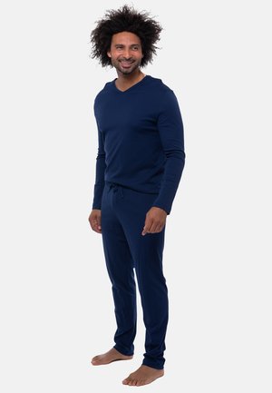 Man met krullend haar, gekleed in een marineblauw shirt met lange mouwen en broek, staat op blote voeten en glimlacht, terwijl hij opzij kijkt.